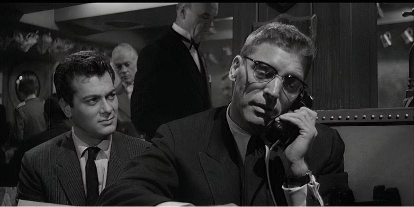A man wearing glasses talks on a landline phone while another man watches in Sweet Smell of Success.