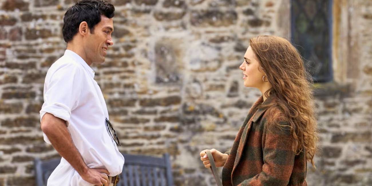 Alex Hassell and Bella Maclean talk, facing each other in front of a brick building in Rivals Season 1. 
