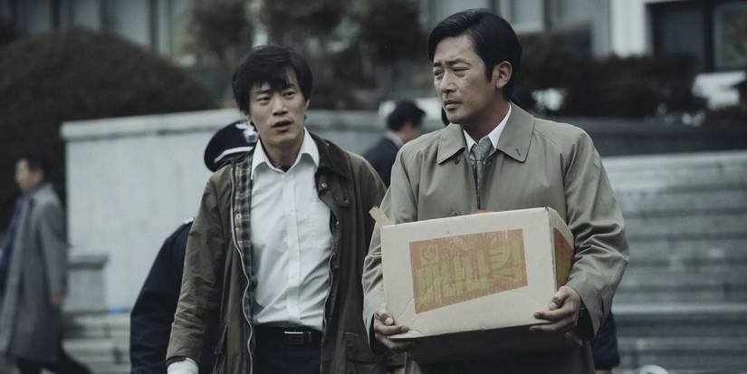 Two men walking on the street and talking; one of them is holding a box labeled in Korean language, and the other is behind him, telling him something