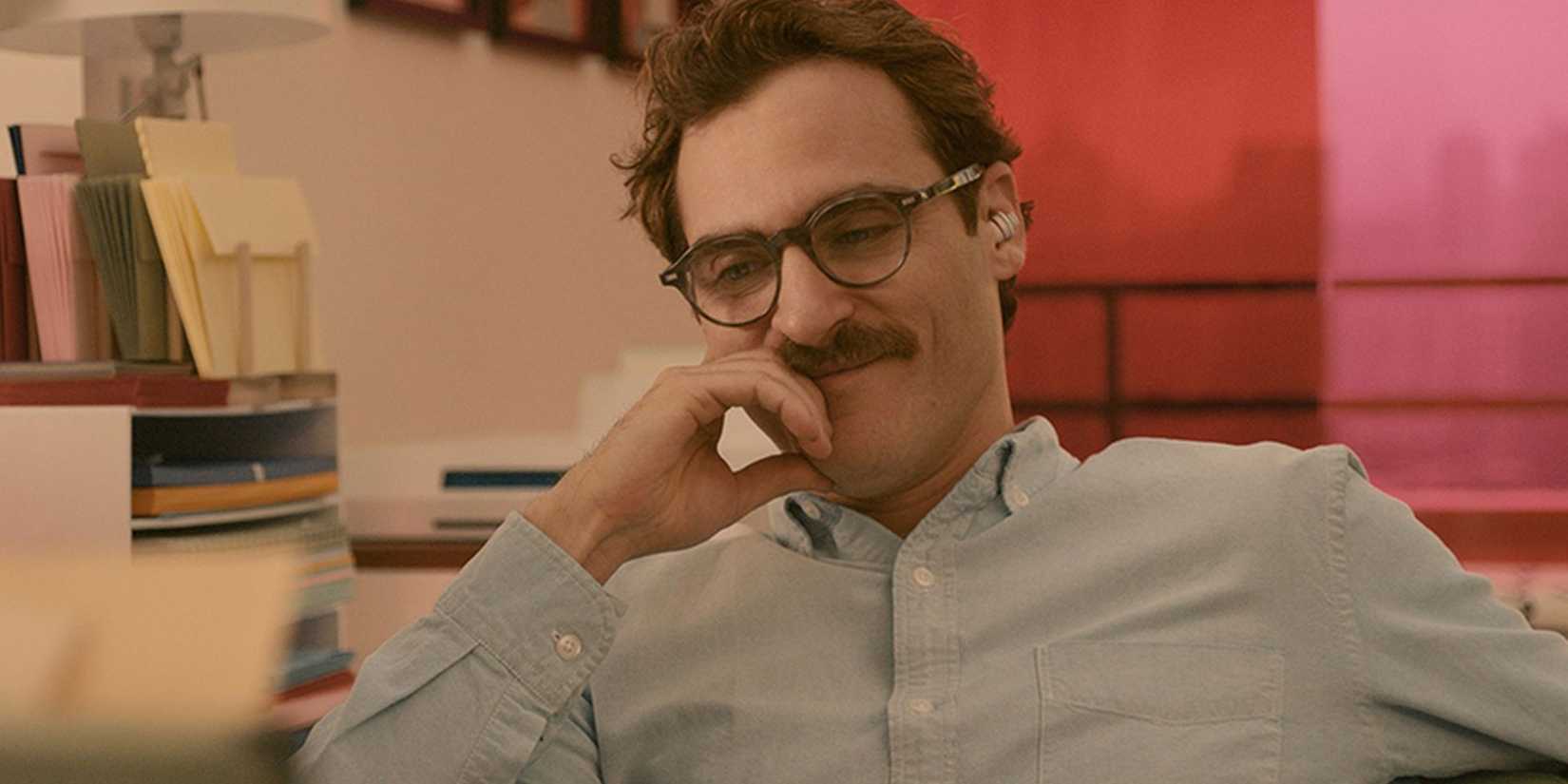 Joaquin Phoenix sitting at a desk with a hand to his mouth & smiling in HER