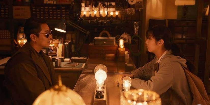 A man and woman sit across from each other at a table with light bulbs all around in Light Shop.