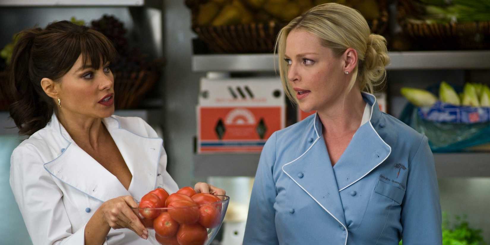 Sofia Vergara holds a bowl of tomatoes in a restaurant kitchen while Kathryn Heigl scowls