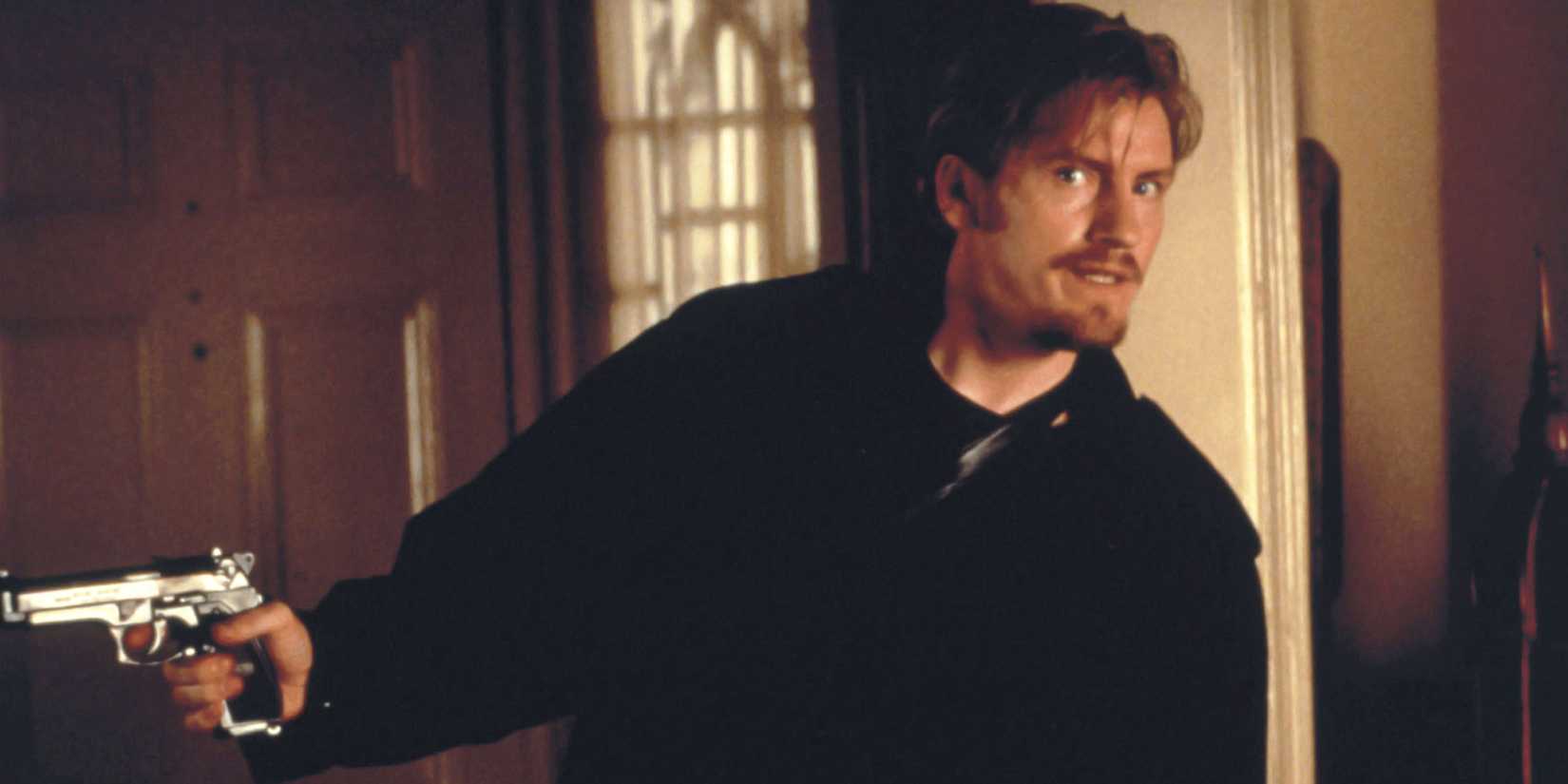 Denis Leary dressed in black pointing a gun off-camera in The Ref - 1994