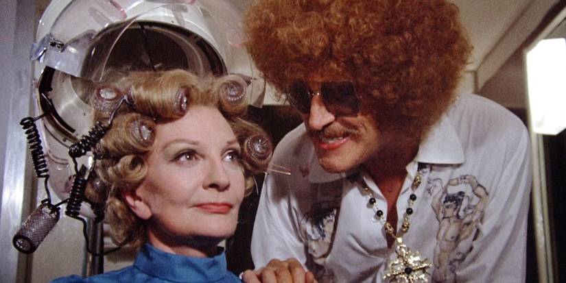 Edward Lionheart (Vincent Price) mengenakan afro coklat dan baju olahraga putih berkilauan tahun 70-an, bersandar di bahu korban perempuan (Coral Browne) saat dia duduk di bawah pengering salon rambut di Theater of Blood