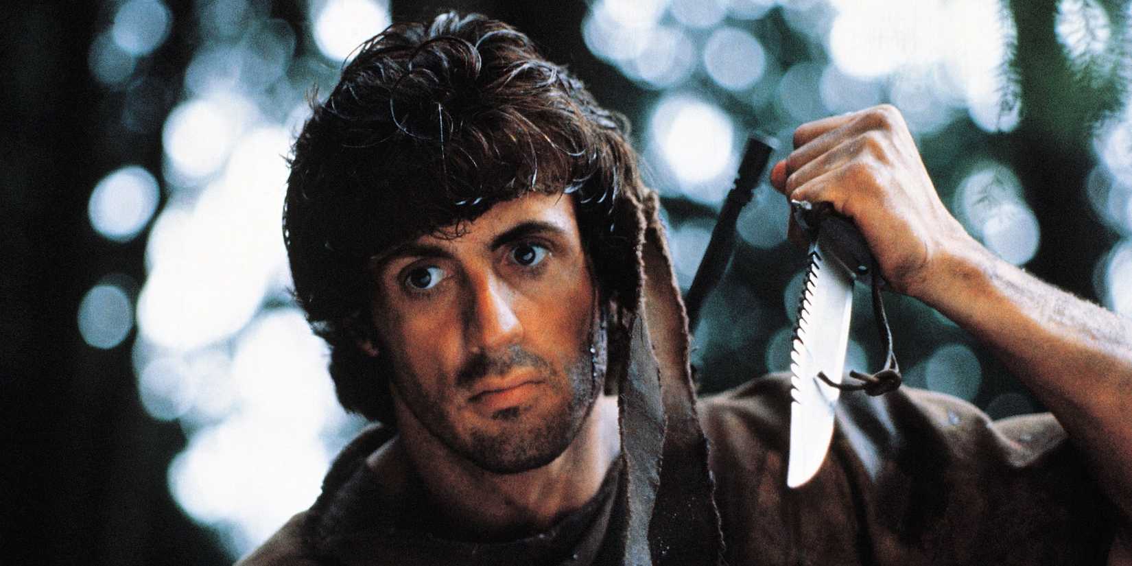 Actor Sylvester Stallone as Rambo, looking down at something and raising a knife above him in First Blood.
