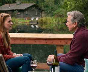 Mel and Doc sitting on lounge chairs on a patio in front of a wooden cabin and a lake in Virgin River.