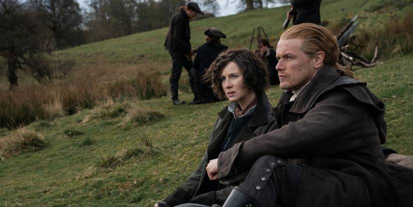 Caitriona Balfe as Claire and Sam Heughan as Jamie sit on a hillside in period clothes in Outlander.