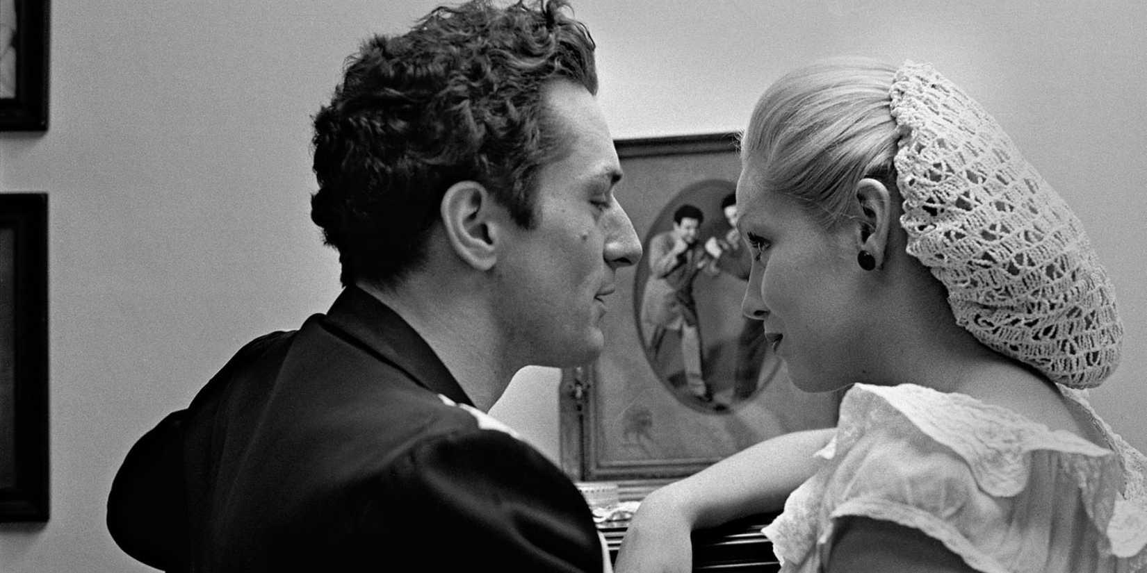 A man talking to a woman who is wearing a snood as they stand near a picture in Raging Bull.