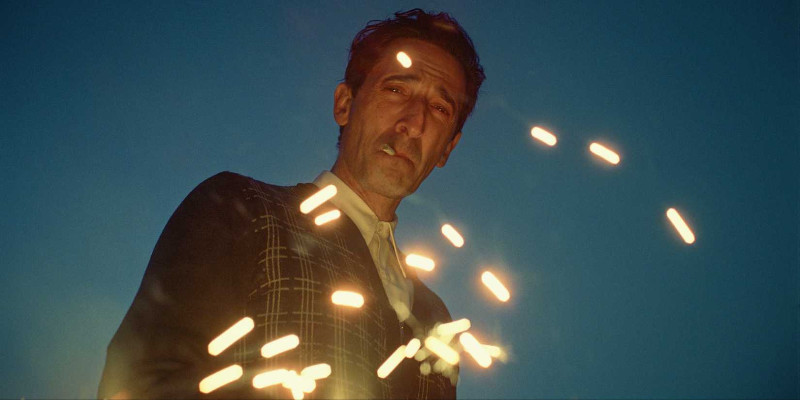 Adrien Brody staring at the ground at dusk while sparks fly around him in 'The Brutalist'