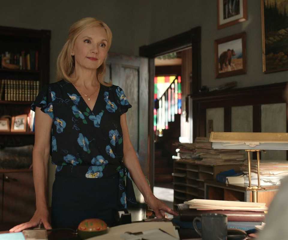 A woman stands and talks to a man who is sitting behind a desk in a home office in Virgin River.