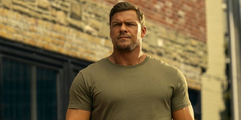 Alan Ritchson as Jack Reacher, in a beige t-shirt, standing in front of a brick building in Reacher Season 3.