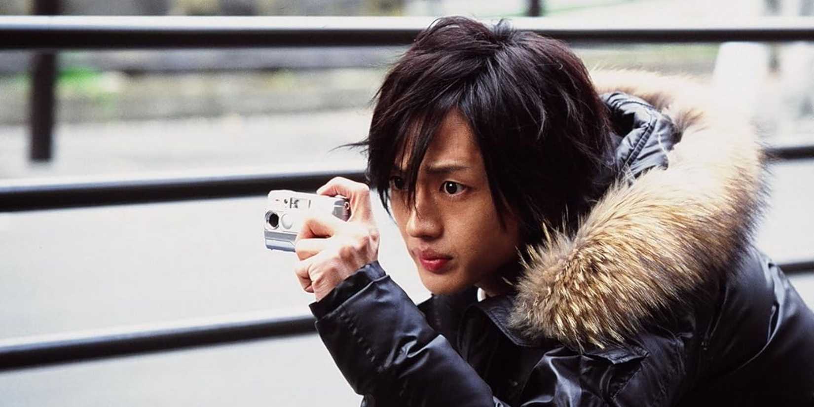 A man stands in a winter jacket aiming a camera in Love Exposure.