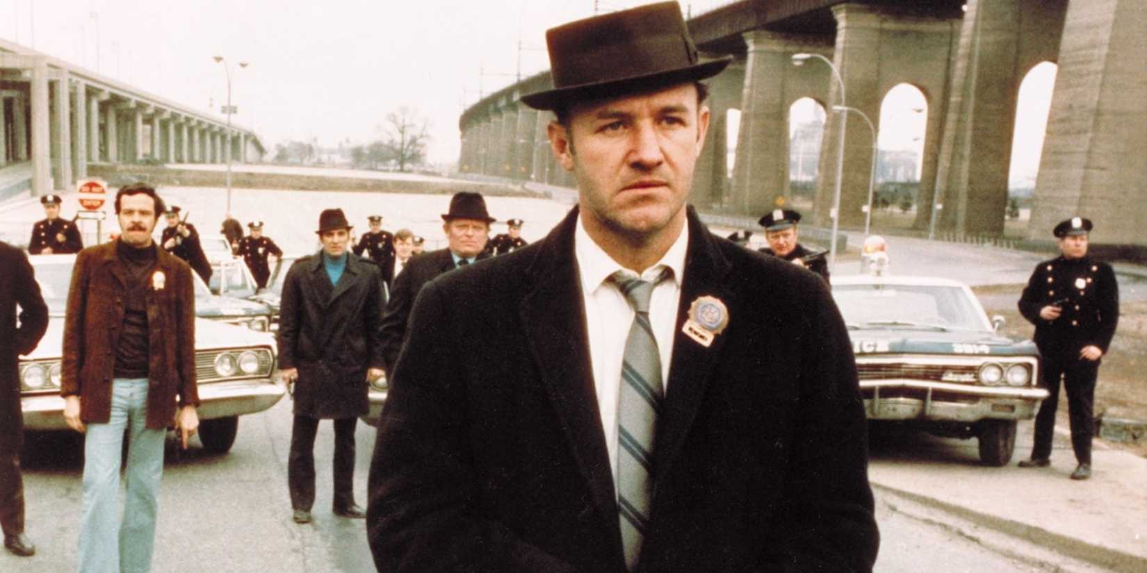 Gene Hackman as Popeye Doyle standing in a street with officers behind in The French Connection.