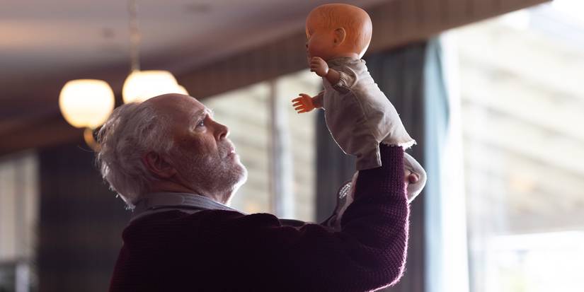 John Lithgow holding up a toy doll and looking up at it sadly