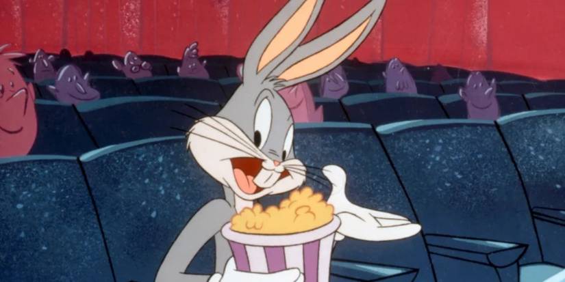 Bugs Bunny sits in a movie theater audience smiling at the camera and holding popcorn in Looney Tunes.