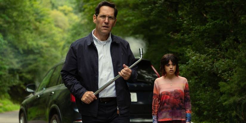 Paul Rudd holds a crowbar in front of Jenna Ortega, both walking toward something in the road in Death of a Unicorn.