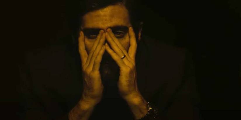 Jake Gyllenhaal sits in a dark room and uses his hands for an odd pose in the thriller Enemy.
