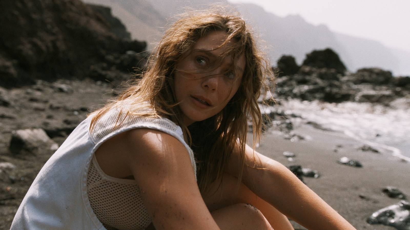 Elizbeth Olsen as Mia sitting in the sand on the beach and looking over her right shoulder in The Assessment