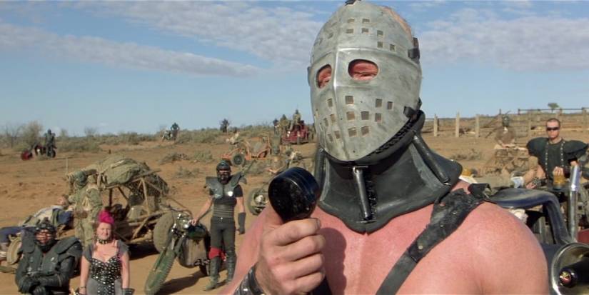 A person with a metal covering over their face with others and makeshift cars in Mad Max 2 The Road Warrior. 