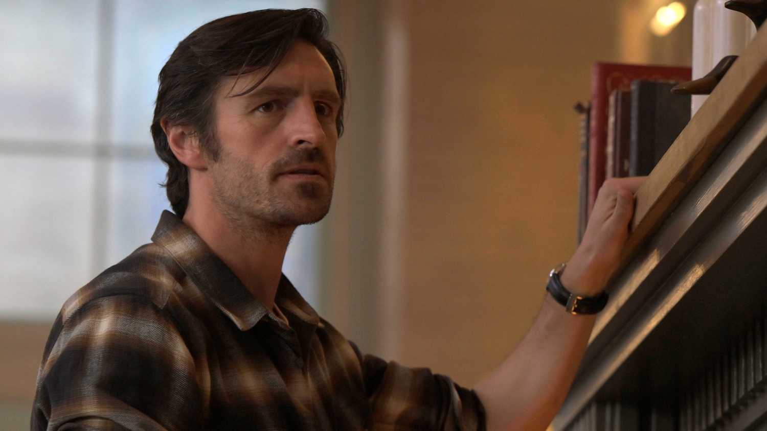 Eoin Macken as Davis Collins with his left hand up on the mantle in a plaid shirt in Ransom Canyon