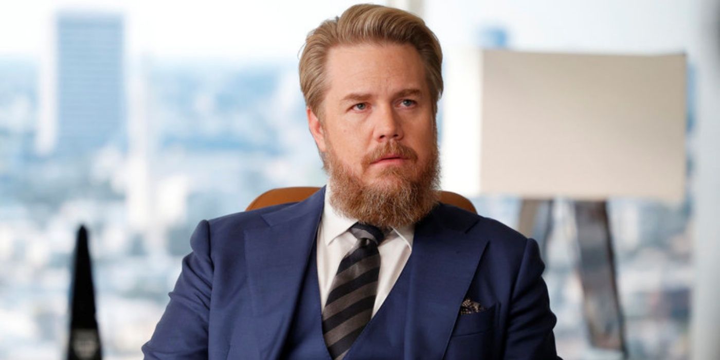 Josh McDermitt as Stuart Lane sitting in a chair in his office while wearing a suit in Suits LA