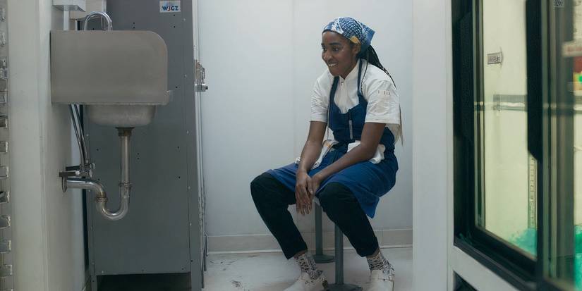 Sydney (Ayo Edebiri) sits on a stool next to lockers and contemplates quitting her job in The Bear on Hulu.