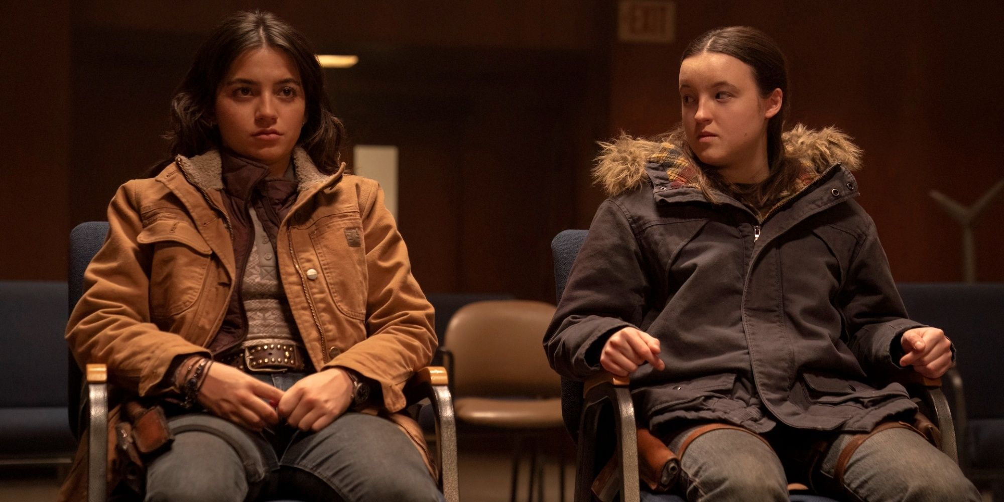 Isabela Merced as Dina sitting in a chair next to Bella Ramsey as Ellie in The Last of Us Season 2