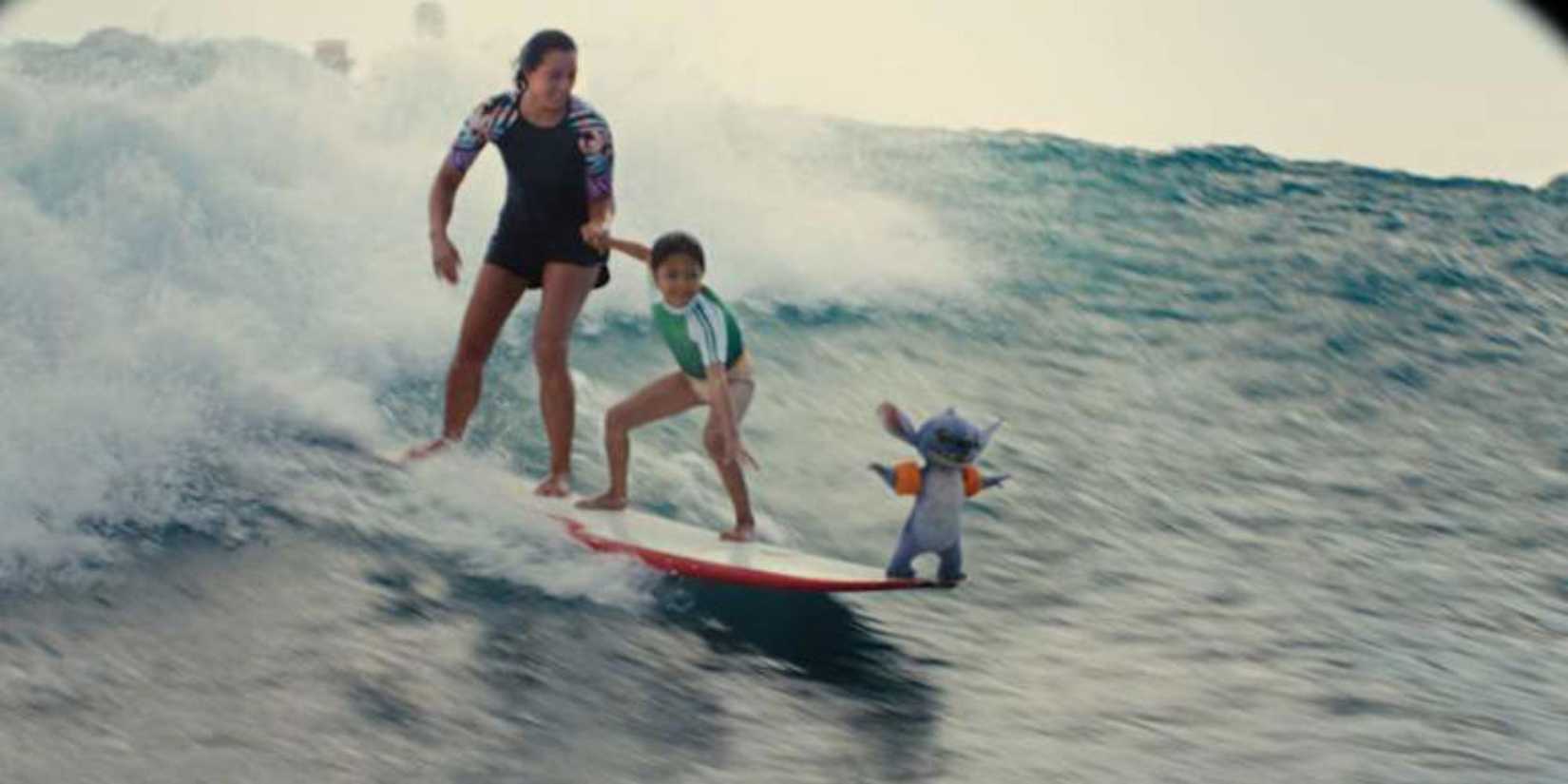 Sydney Agudong as Nani and Maia Kealoha as Lilo, surfing while Stitch stands on the front of their board in Lilo & Stitch.