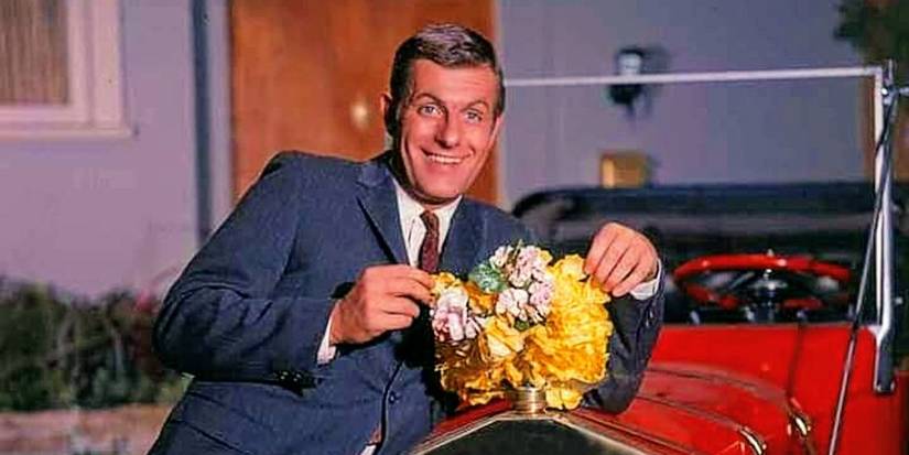 Dave standing in front of a red car holding flowers next to it in My Mother the Car.