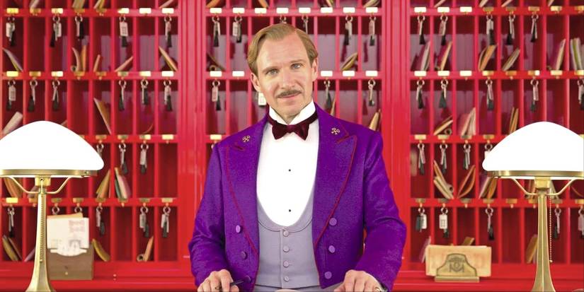 Ralph Fiennes as M. Gustave in a purple tuxedo stands behind the front desk in The Grand Budapest Hotel.