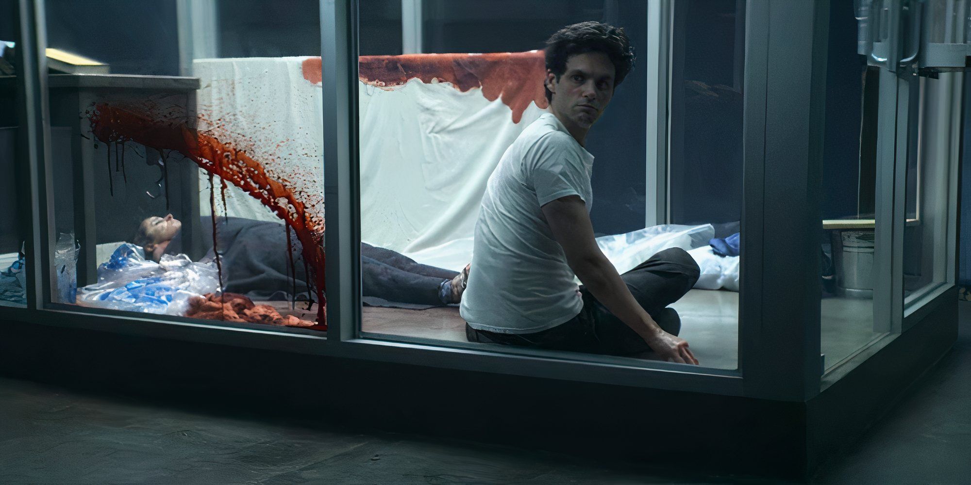 Penn Badgley as Joe in You's P.I. Joe sits in a glass room next to a bloody body and looks over his shoulder.