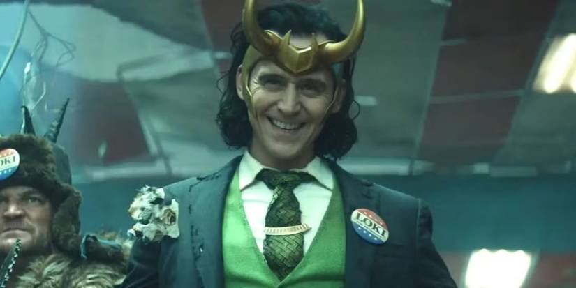 Tom Hiddleston as President Loki smiling mischievously with a tattered suit and gold horns in Loki.