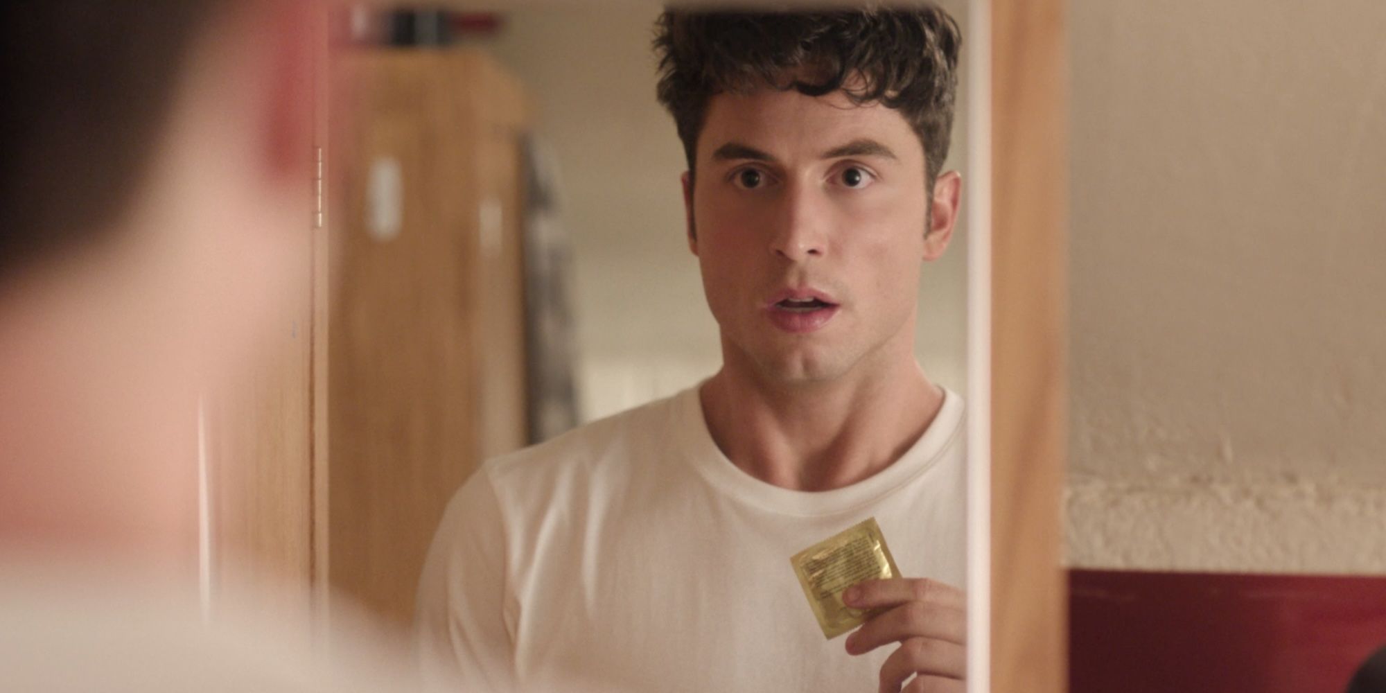 Benito Skinner as Benny looking in the mirror holding a condom in Overcompensating