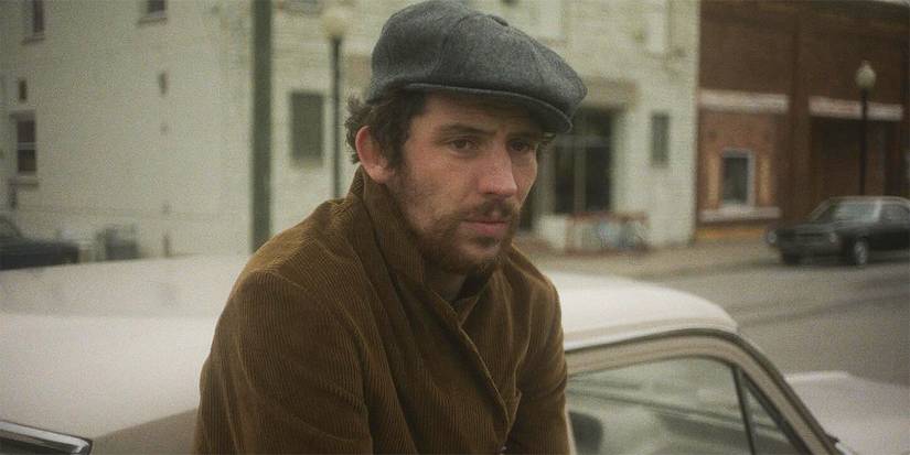 Josh O'Connor wearing a coat and cap while leaning against a car in The Mastermind
