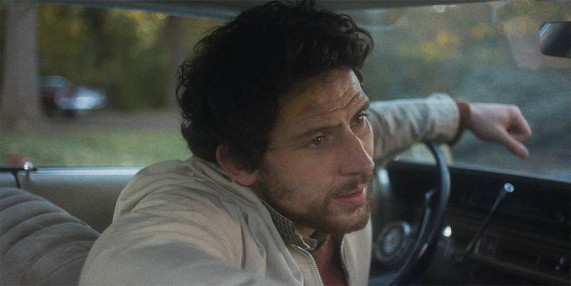 Josh O'Connor sitting in a car and talking to someone with a bandage on his forehead in The Mastermind.