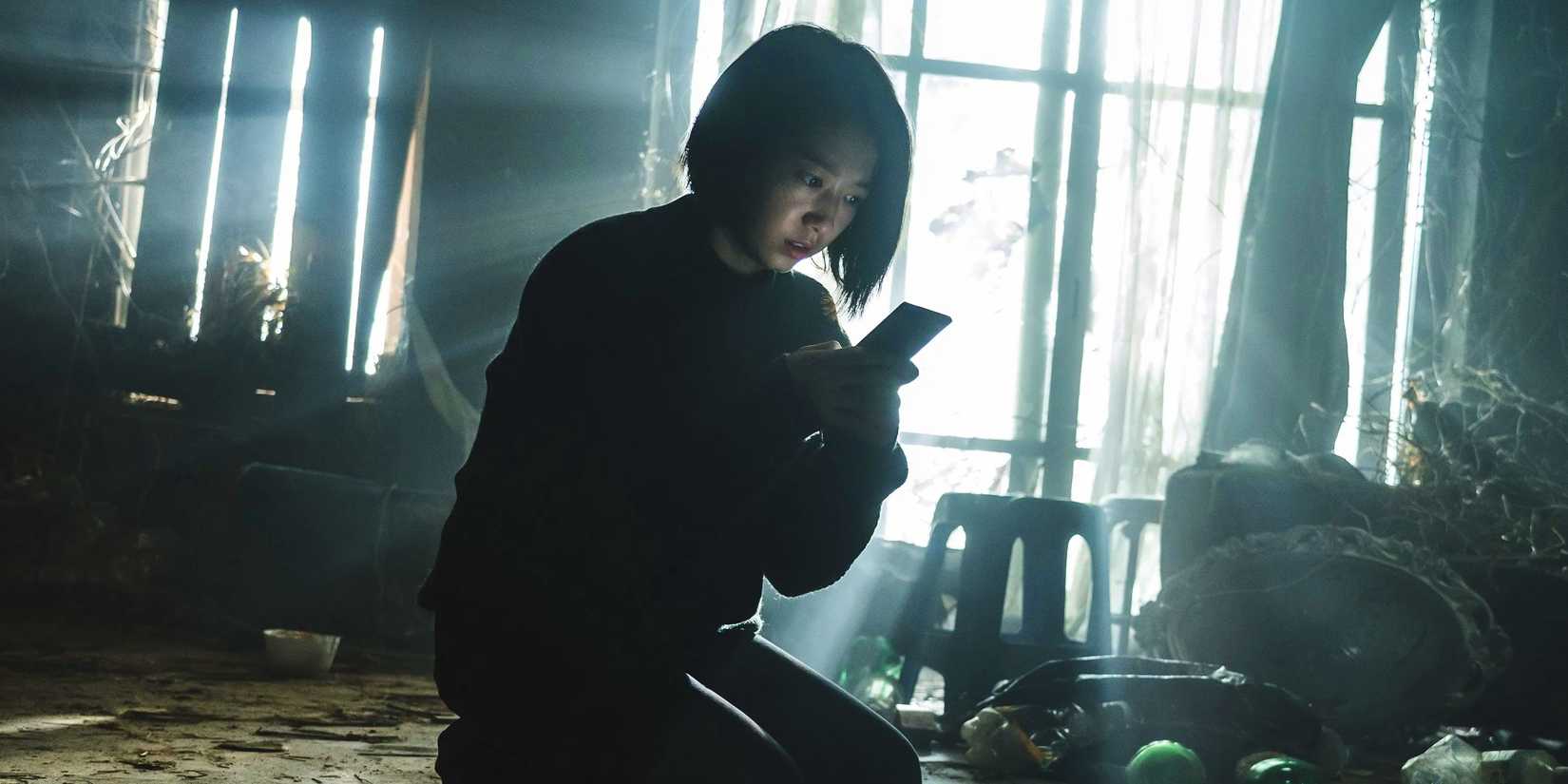 Actor Park Shin-hye as Seo-yeon, knelt in a dark, ramshackle house, looking worriedly at her cell phone in The Call.