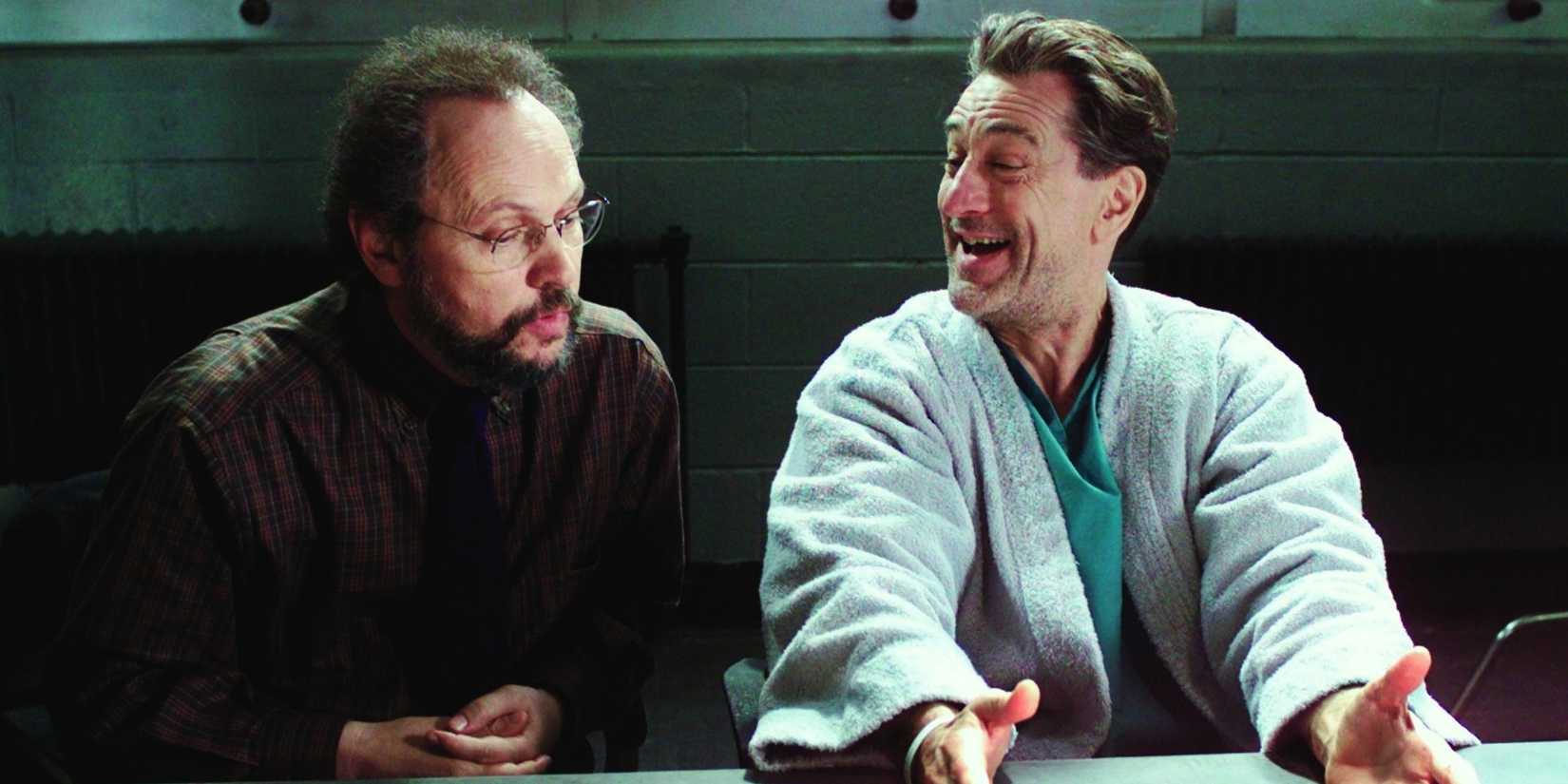 Paul, played by actor Robert De Niro, sits at a table in a bathrobe smiling and gesturing with his hands while Ben, played by actor Billy Crystal, sits next to him straight-faced in Analyze This.