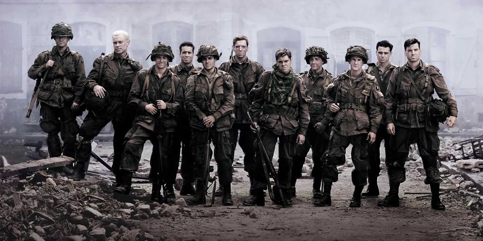 The cast of Band of Brothers stand in a row amidst rubble, looking at the camera wearing military uniforms. 