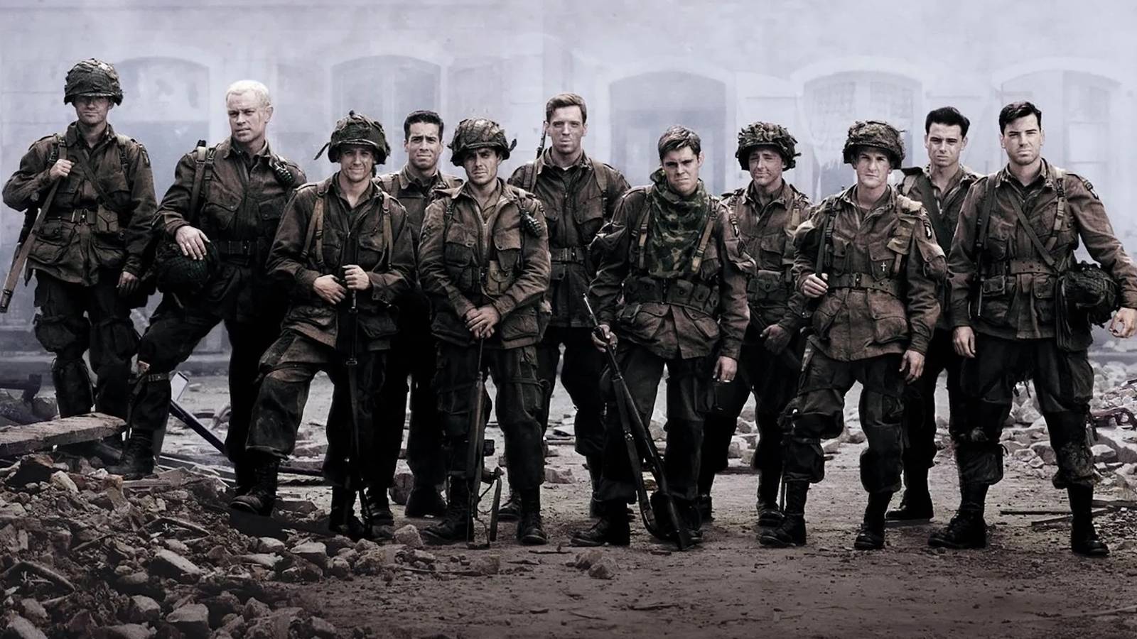 The cast of Band of Brothers stand in a row amidst rubble, looking at the camera wearing military uniforms. 