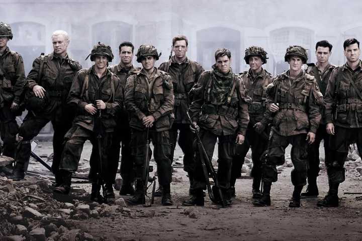The cast of Band of Brothers stand in a row amidst rubble, looking at the camera wearing military uniforms. 