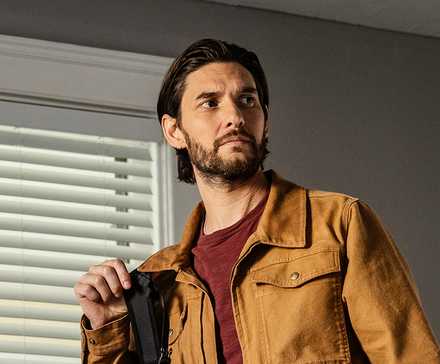 Ben Barnes as Tim standing inside a house next to a window with blinds in The Institute