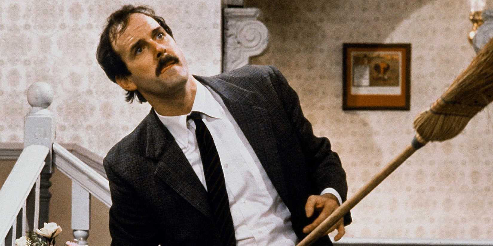 John Cleese as Basil Fawlty holding a broom to a wall and looking up in Fawlty Towers.