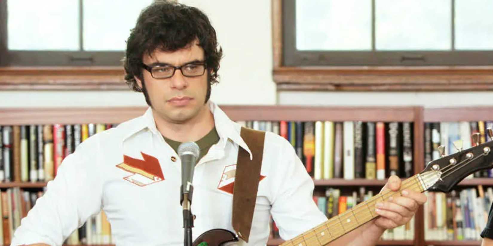 Jemaine holding a guitar and standing in front of a microphone in a library in Flight of the Conchords.