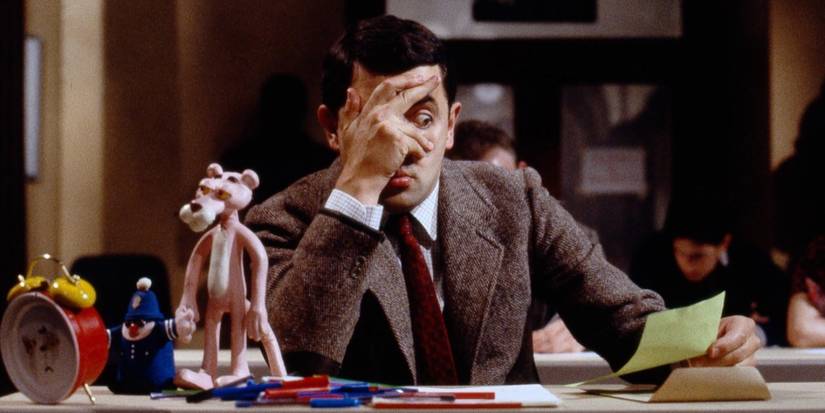 Rowan Atkinson as Mr. Bean covering his face as he peers at a piece of paper in his hand in Mr. Bean, 1990.