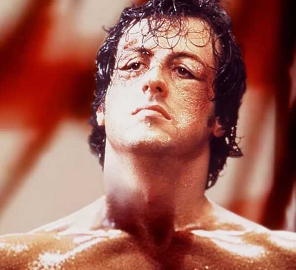 Actor Sylvester Stallone as Rocky Balboa in Rocky, looking exhausted but defiant in front of an American flag.