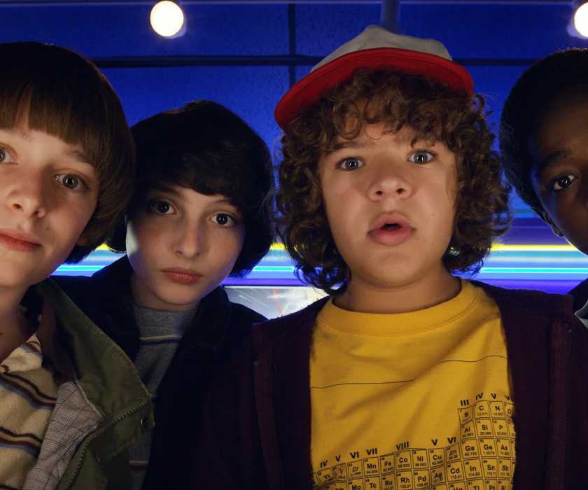 Will, Mike, Dustin, and Lucas watching an arcade game in Stranger Things.