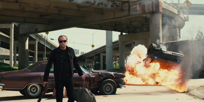 A man in shades, carrying a shotgun and a bag walks away from a car explosion