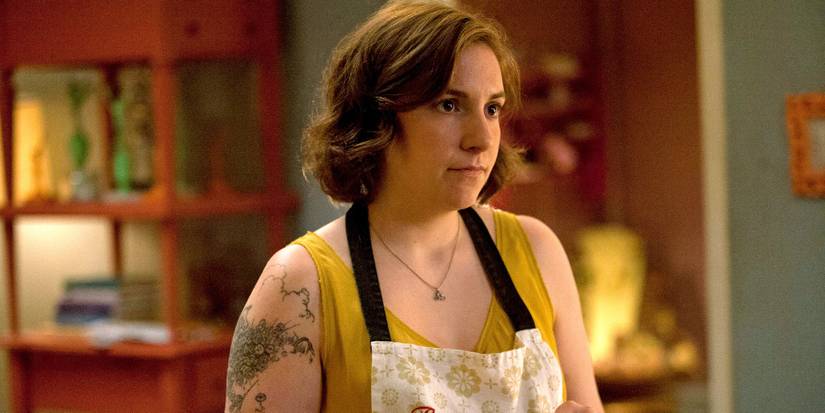 Hannah Horvath (Lena Dunham) stands in her house wearing a yellow shirt and an apron in 'Girls' (2012-2017).