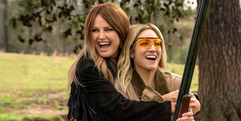 Malin Åkerman as Margo hugs Brittany Snow as Sophie from behind while holding a rifle in 'The Hunting Wives.'