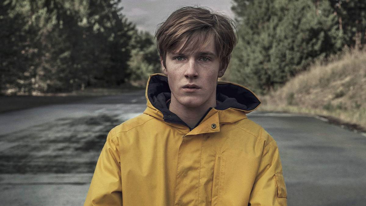 Louis Hofmann in a yellow raincoat standing on a deserted road in Dark.
